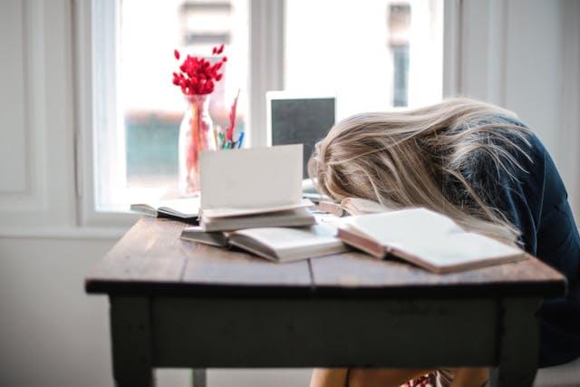 Femme blonde aux cheveux longs qui dort que une bureau en bois avec des livres ouverts et un vase avec des fleurs rouges